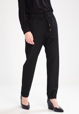 Frau trägt schwarze, zulaufende Hose mit Kordelzug und schwarze High Heels, stehend vor weißem Hintergrund.