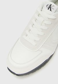 Witte Calvin Klein sneaker met suède en leren details, witte veters en een zwarte zool met de tekst "Calvin Klein Jeans".