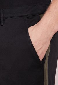 Black pants with a side pocket in a contrasting olive green panel. The fabric appears smooth, with visible stitching details along the waistband.