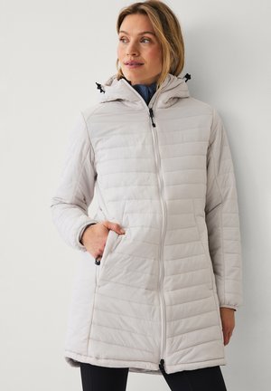 Vrouw draagt een lichtbeige doorgestikte jas met capuchon, voorzien van een ritssluiting aan de voorkant en zakken, staand tegen een effen lichte achtergrond.