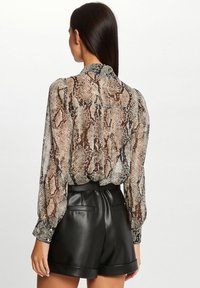 Transparante blouse met een bruin en zwart slangenprint, lange mouwen, hoge halslijn en elastische zoom, gecombineerd met zwarte leren shorts.
