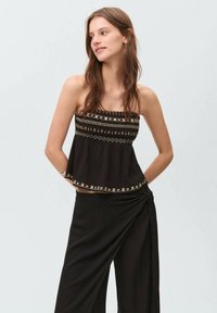 Svart bandeau-topp med broderade gyllene geometriska mönster längs halsringningen och fållen, i kombination med flödande svarta byxor. Mjuk textur.