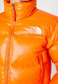 Pomarańczowa, lekka kurtka puchowa z wysokim kołnierzem, z centralnym zamkiem błyskawicznym i białym logo na piersi, wykonana z błyszczącego, gładkiego materiału.