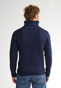 Donkerblauwe gebreide trui met een hoge geribbelde kraag, textuurstof met subtiele spikkels, aansluitend bij de taille en polsen, gecombineerd met jeans.