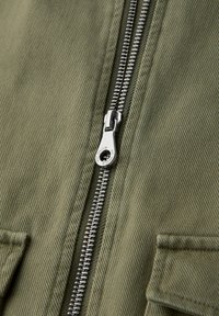 Nahaufnahme eines teilweise geschlossenen silbernen Metallreißverschlusses auf olivgrünem, strukturiertem Stoff mit sichtbaren Nähten und zwei Stofflaschen.