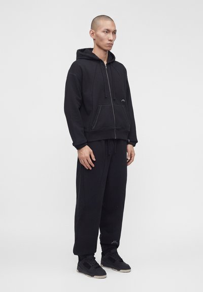 Άνδρας που στέκεται φορώντας μαύρο φούτερ με φερμουάρ, αντίστοιχο φόρμας και μαύρα αθλητικά παπούτσια μπροστά σε απλό γκρι φόντο.