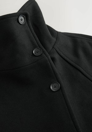 Melna jaka ar augstu apkakli, diviem melniem pogām un teksturētu audumu ar redzamām šuvēm.