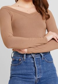 Långärmad, figursydd topp i ljusbrunt tyg med V-ringning. Kombinerad med högmidjade blå denimjeans med klassiska fickdetaljer.