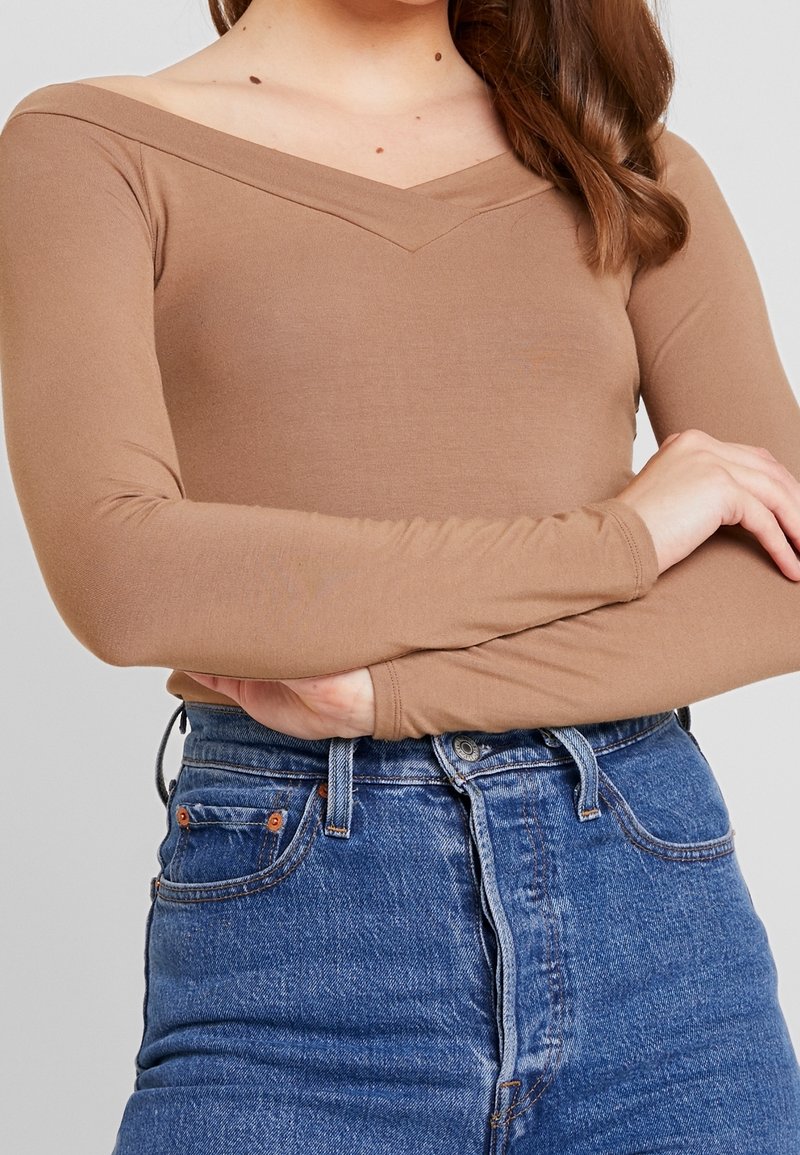 Långärmad, figursydd topp i ljusbrunt tyg med V-ringning. Kombinerad med högmidjade blå denimjeans med klassiska fickdetaljer.