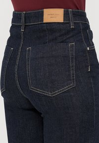 Mørkeblå denim jeans med synlig kremfarget søm, baklomme og en brun lærmerkelapp som sier "perfect jeans gina tricot."