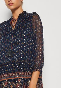 Marinblå blommig klänning med volangkrage, långa genomskinliga ärmar och en vågig nederkant. Har ett mönstrat band vid midjan och franstillbehör.