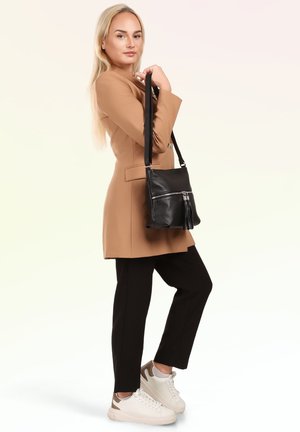 Blonde vrouw in een beige jas, zwarte broek en witte sneakers die een zwarte leren schoudertas met rits en sierkwastjes vasthoudt.