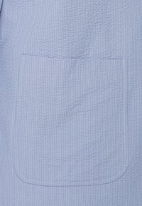 Tela azul claro texturizada con un patrón de cuadrícula, que presenta un gran bolsillo parche cosido con hilo blanco.