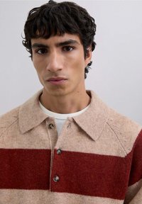Suéter de punto con base beige y dos franjas rojas anchas, que cuenta con un cuello, una tapeta abotonada y una tela texturizada.