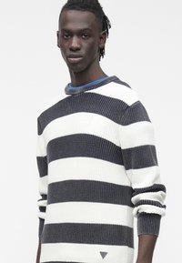 Mlad moški s temno poltjo in kratkimi dreadlocki nosi temno modro in belo črtasto pleteno pulover, stoji pred preprostim belim ozadjem.