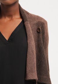 Brauner Wollcardigan mit weicher Textur, offener Vorderseite und einem einzigen braunen Knopf am Kragen, kombiniert mit einem schwarzen V-Ausschnitt-Oberteil.