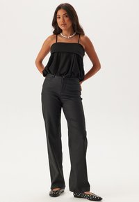 Top nero senza maniche con un design a strati, abbinato a pantaloni neri. Il modello indossa ballerine nere con pois bianchi e una collana di perle.