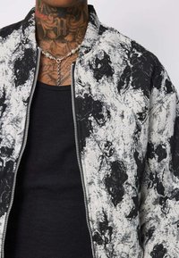 Veste bombardier noire et blanche avec un motif floral texturé, dotée d'une fermeture éclair argentée et d'un ourlet côtelé, portée par-dessus un débardeur noir.