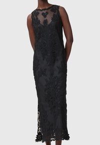 Robe midi noire sans manches avec haut transparent et broderie florale en dentelle portée par une personne à la peau foncée, debout les mains derrière le dos.