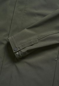 Détail de la manche et du poignet d'une veste vert foncé avec fermeture par bouton-pression et coutures visibles.