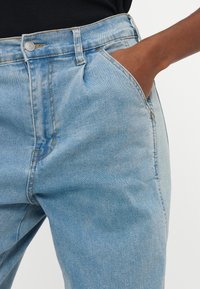 Δεξί χέρι στην τσέπη τζιν παντελονιού από ανοιχτό μπλε ντένιμ, φορεμένο με μαύρο τοπ, ορατή μέση και ραφές στο τζιν.