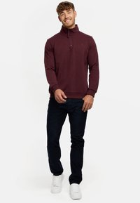 Burgunderfarbener Zip-Hoodie mit hohem Kragen, langen Ärmeln und gerippten Bündchen, kombiniert mit dunklen Jeans und weißen Turnschuhen.