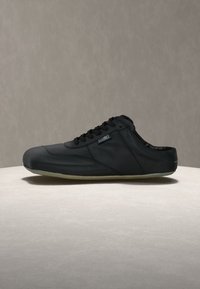 Μαύρο καθημερινό παπούτσι με κορδόνια και μαλακή, ευέλικτη σόλα, τοποθετημένο σε στρογγυλή μπεζ επιφάνεια με φόντο ένα απλό γκρι φόντο.