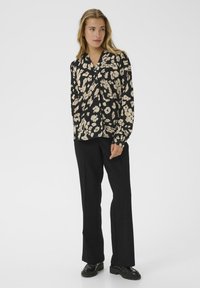 Zwarte bloes met beige abstracte patronen, lange mouwen, V-hals en knoopsluiting. Gecombineerd met zwarte rechte broek en zwarte schoenen.