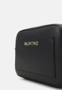 Bolso de cuero negro con un acabado texturizado, que presenta un logo dorado de "VALENTINO" y una forma rectangular elegante. Sin patrones decorativos.