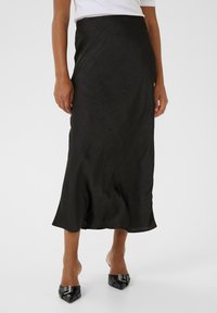 Jupe midi en satin noir avec un ourlet légèrement évas�é et une texture subtile, associée à des talons noirs pointus avec des éléments décoratifs.