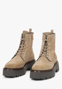 Beżowe zamszowe botki za kostkę z perforowanymi detalami, brązowa platforma, sznurowany przód i zamek błyskawiczny z boku. Teksturowane wykończenie z pętelką.