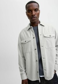 Camisa de botón gris claro hecha de una tela suave y texturizada, con dos bolsillos de solapa y botones oscuros, superpuesta a un suéter de punto gris oscuro.