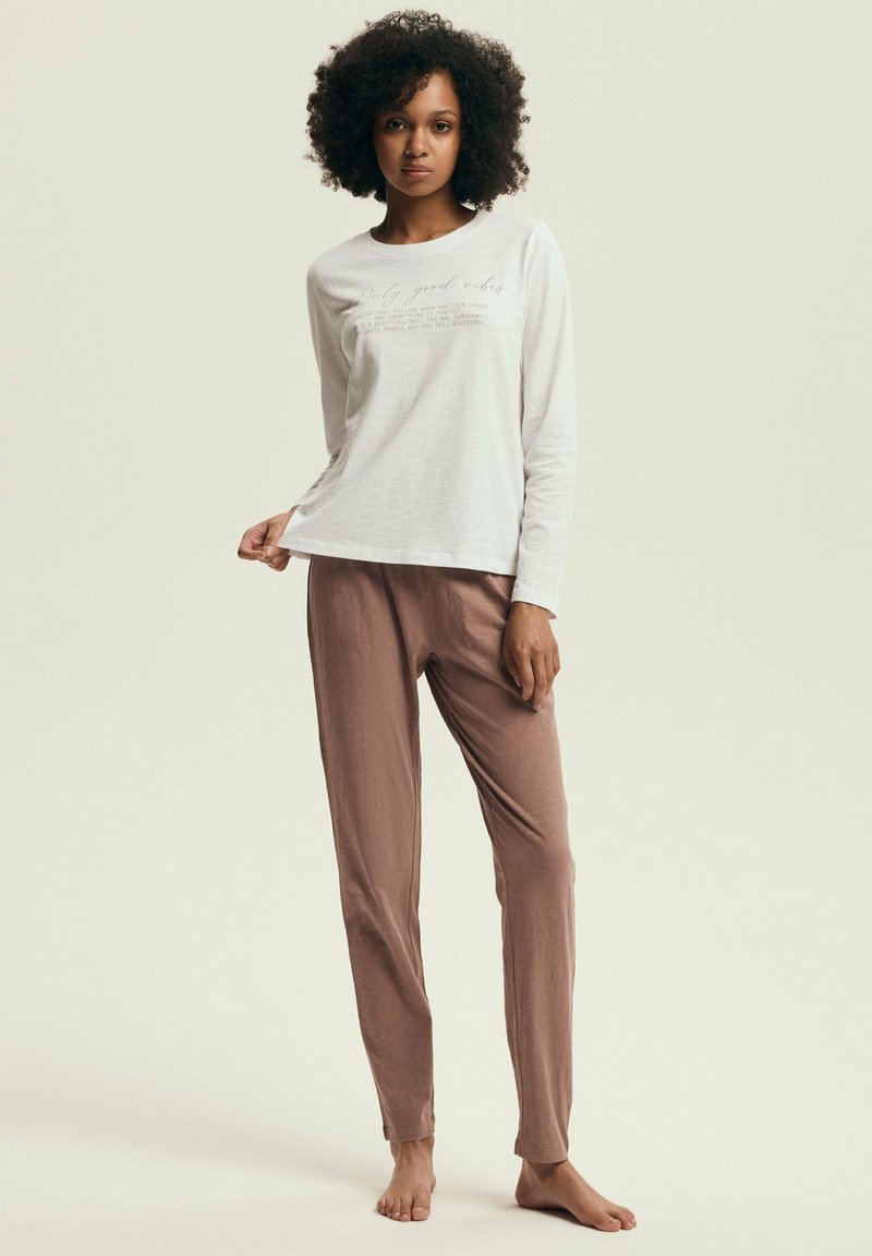 Biała bluzka z długim rękawem z nadrukiem, zestawiona z miękkimi, jasnobrązowymi, luźnymi spodniami. Materiał wydaje się gładki i lekki.