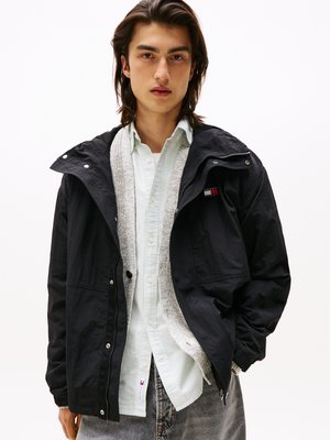 Giacca nera con zip frontale, rifinita con un morbido rivestimento grigio e una piccola patch con logo; indossata sopra una camicia a righe verde chiaro.