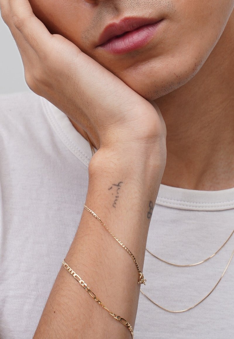 Gold-tone bracelets on a wrist with a tattoo, against a background of a white t-shirt. Skin texture is visible and subtle.