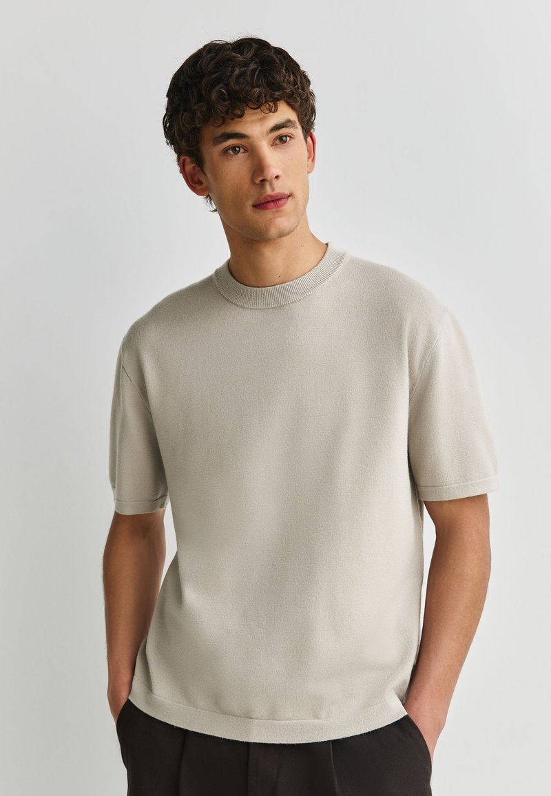 Giovane uomo con capelli ricci e scuri che indossa una maglietta beige chiaro a maniche corte in maglia e pantaloni neri, con le mani nelle tasche, che guarda leggermente di lato.