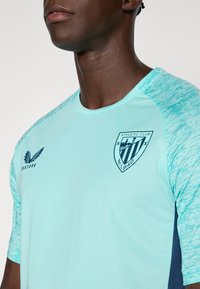 Mężczyzna w jasnoniebieskiej koszulce piłkarskiej Athletic Club Bilbao z teksturowanymi rękawami i logo klubu na piersi.