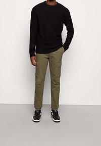 Homme portant un pull en tricot noir, un pantalon cargo vert olive avec des poches boutonnées, et des baskets noires et blanches, debout devant un fond uni.