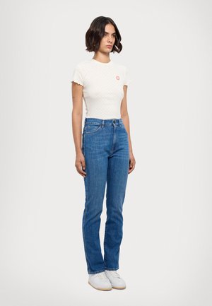 Witte short-sleeve shirt met golvende randen en een klein rood logo, gecombineerd met blauwe straight-leg jeans en witte sneakers.
