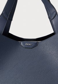 Τσάντα χειρός από δέρμα σε ναυτικό μπλε με υφασμάτινη επιφάνεια, διαθέτει λεπτή λαβή και ασημί λογότυπο στο μπροστινό μέρος.