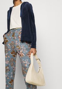 Henkilö, joka käyttää meripeninkulman väristä vetoketjullista hupparia, valkoista t-paitaa ja sinisiä kukkakuosisia housuja, pitäen kädessään kermaista, teksturoitua käsilaukkua, jossa on lyhyt hihna.