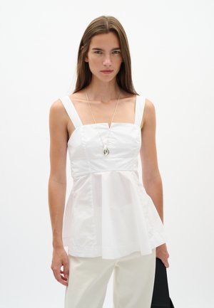 Jeune femme aux longs cheveux bruns porte un haut blanc sans manches, un pantalon blanc, un collier pendentif en argent et tient un sac noir sur fond blanc.