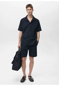 Cămașă cu mânecă scurtă, de un albastru închis, cu nasturi, pantaloni scurți asortați și sandale negre. Accesoriile includ o geantă structurată de culoare închisă, cu mânere.