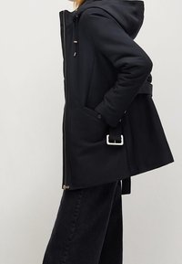 Parka noire à capuche avec fermeture éclair, poches avant, ceinture avec boucle blanche et finition mate lisse. Longueur mi-cuisse.