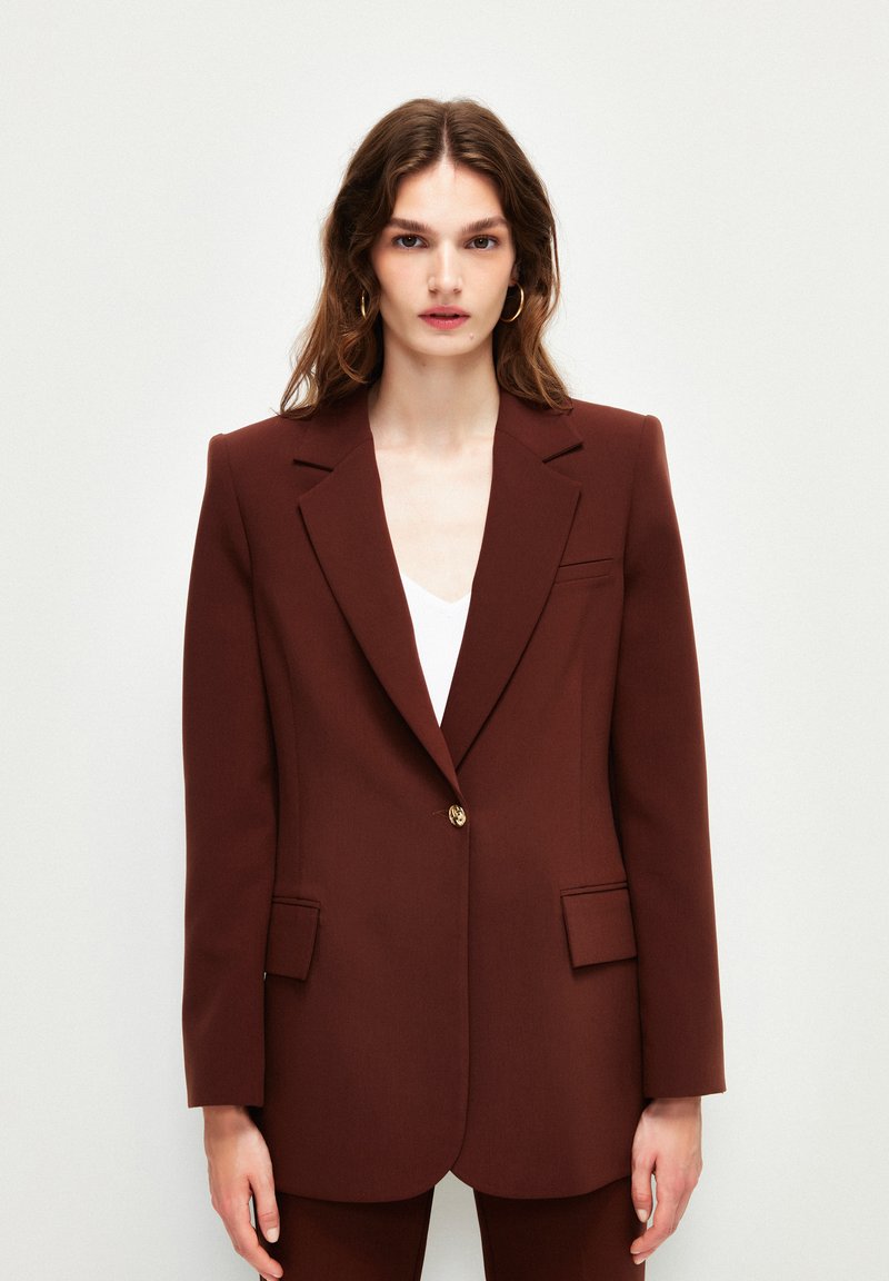 Bruine blazer met een enkele gouden knoop, scherpe revers en twee voorzakken. Glad materiaal en getailleerde pasvorm.
