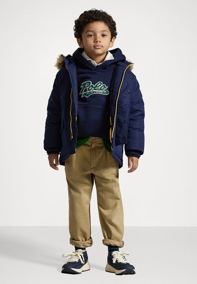 Ναυτική πούφα τζάκετ με κουκούλα και ψεύτικη γούνα, φορεμένη πάνω από ναυτικό φούτερ με πράσινο λογότυπο. Μπεζ παντελόνι και σκούρα αθλητικά παπούτσια.