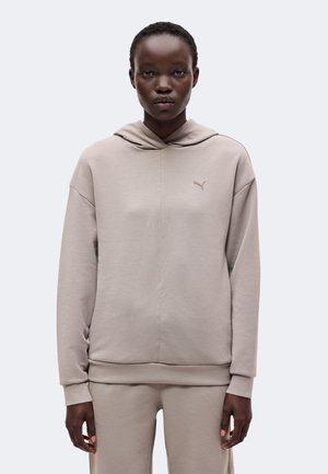 Personne portant un sweat à capuche beige Puma et un pantalon assorti, debout devant un fond clair uni.