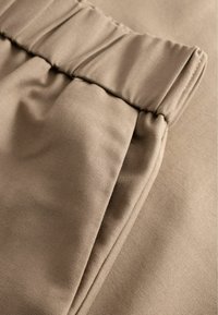 Beżowa tkanina o gładkiej fakturze, z plisami w pasie i miękkim układem tkaniny po bokach, podkreślająca jej strukturę.