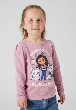 Une jeune fille portant un t-shirt à manches longues rose avec des personnages de chats cartoon et le texte "Les amis de Gabby sont purr-ifiques."