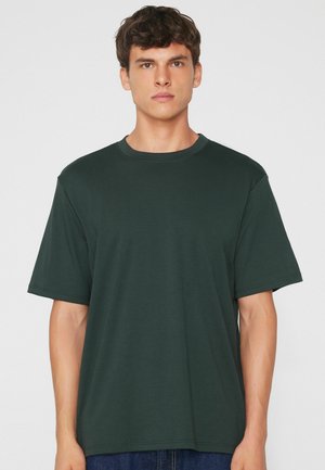 Junger Mann mit kurzen braunen Haaren, der ein dunkelgrünes einfarbiges kurzärmeliges T-Shirt vor einem einfarbigen hellen Hintergrund trägt.